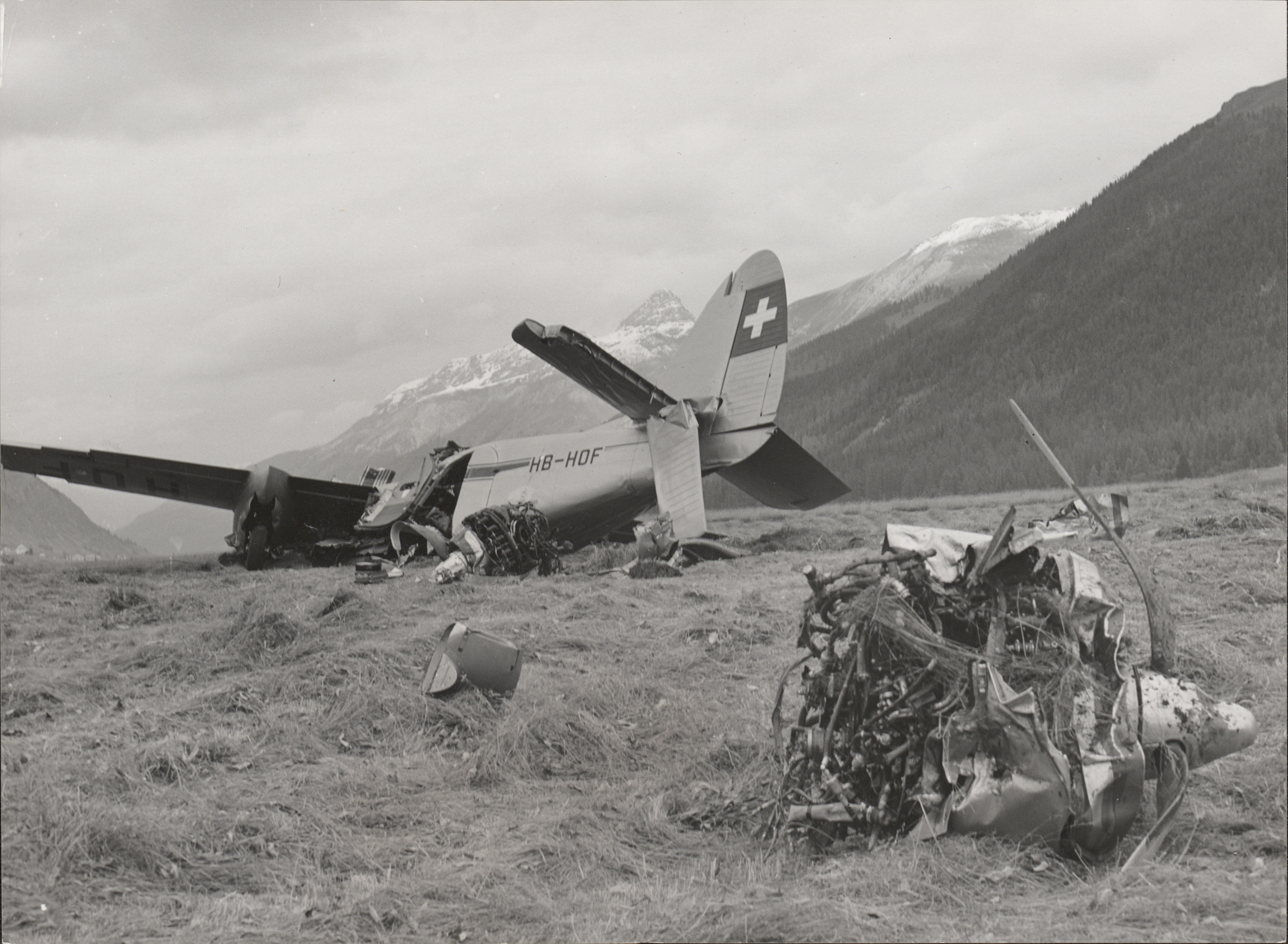 Schwarzweiss-Fotografie eines abgestürzten Flugzeugs vor einer beschneiten Bergkulisse. Im Vordergrund liegen die beiden abgebrochenen Flugzeugmotoren mit verbeulten Propellern.