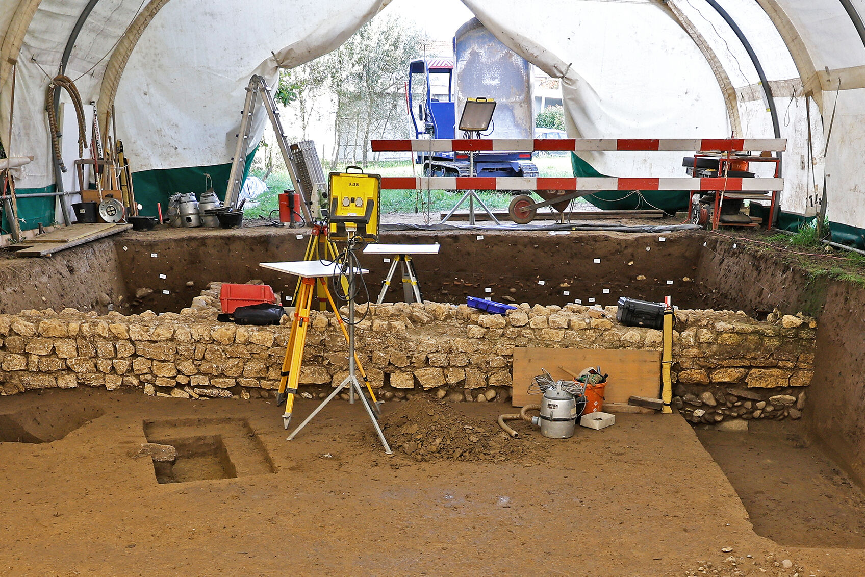 Ausgrabung mit einer römischen Mauer in einem Grabungszelt. Zu sehen sind unterschiedliche Werkzeuge der Archäologie.