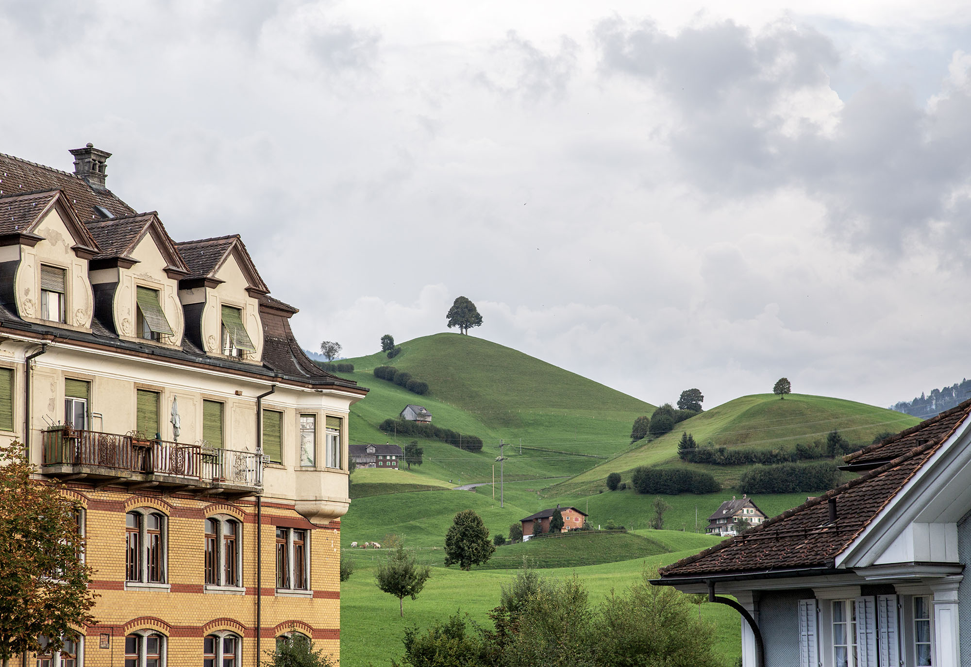 Blick in die Kulturlandschaft Entlebuch, hügelige Landschaft mit bäuerlichen Einzelhöfen. Rechts im Bild die Biskuit-Fabrik, ein origineller Industriebau mit historisierenden Formen und Heimatstilelementen