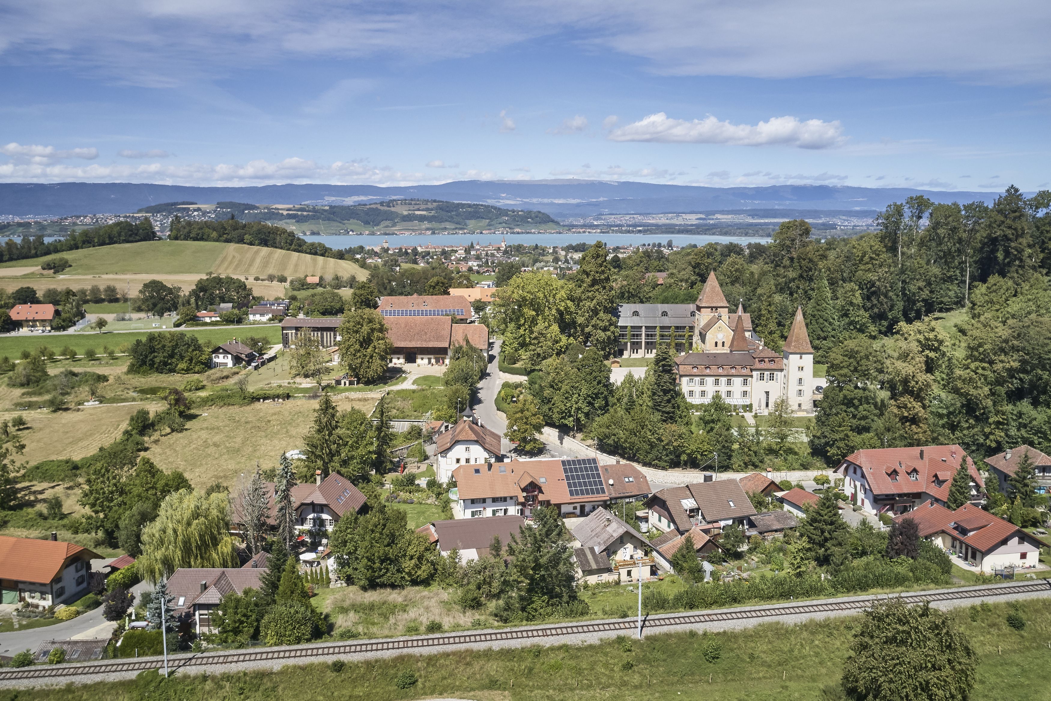 Blick durch die Zieröffnung in der Fassade der alten Voliere zum  Schloss. Aussicht auf das Dorf mit der Burg, dem Hügel Bodenmünzi und dem Murtensee im Hintergrund.